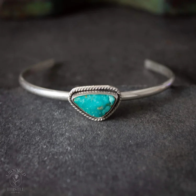 A sterling silver cuff bracelet featuring a triangular turquoise stone centerpiece, resting on a dark textured surface. Crooked Thistle Carico Lake Turquoise Bracelet